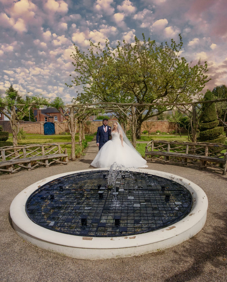 Bride in a white gown and groom in a suit standing on a garden path behind a circular fountain with a tree and wooden benches in the background.