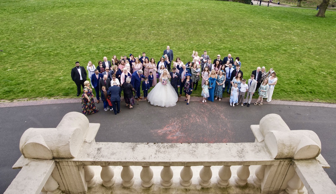 Large group of wedding guests gathered outdoors on a paved area in front of green grass, seen from above with the bride and groom at the centre.