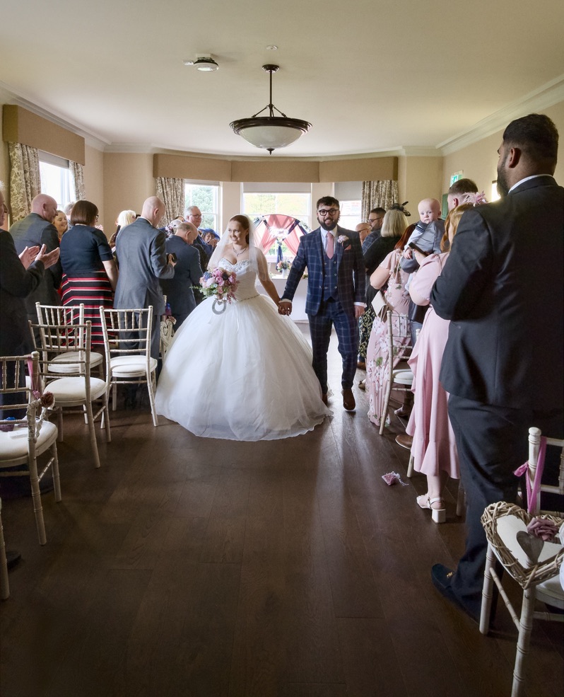 Bride in a white wedding dress and groom in a checked suit walking down the aisle holding hands, surrounded by applauding guests.