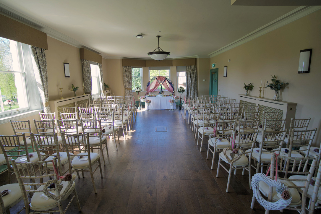 Indoor wedding ceremony setup with rows of white cushioned chairs facing a decorated altar with flowers and draped fabric.