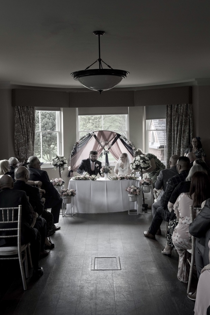 Bride and groom seated at a decorated table during an indoor wedding ceremony with guests seated on either side.