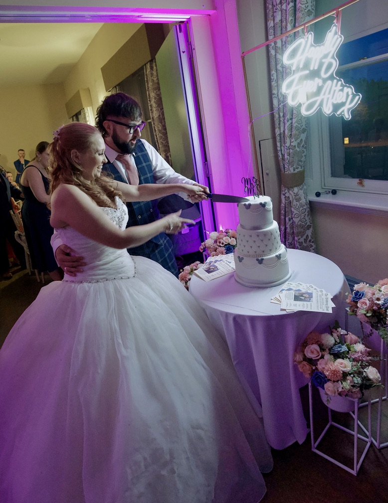 Bride and groom cutting a three-tiered wedding cake at their reception with a glowing 'Happily Ever After' sign in the background.