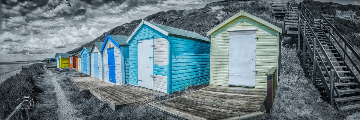 Row of colourful beach huts on a wooden platform beside a sandy shore with stairs leading up a grassy hill under a cloudy sky.