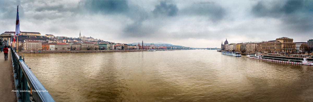 Panoramic view of the Danube River with historic buildings along the riverbank on a cloudy day in Budapest.