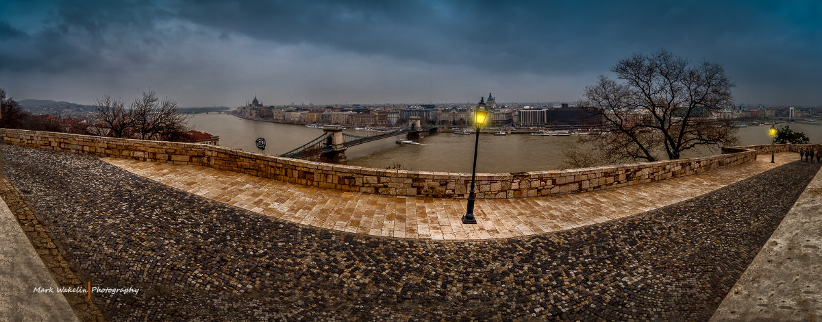 Panoramic view of Budapest riverfront with Chain Bridge, Parliament building, and moody cloudy sky, seen from a stone terrace with cobblestone paths and two lit street lamps.