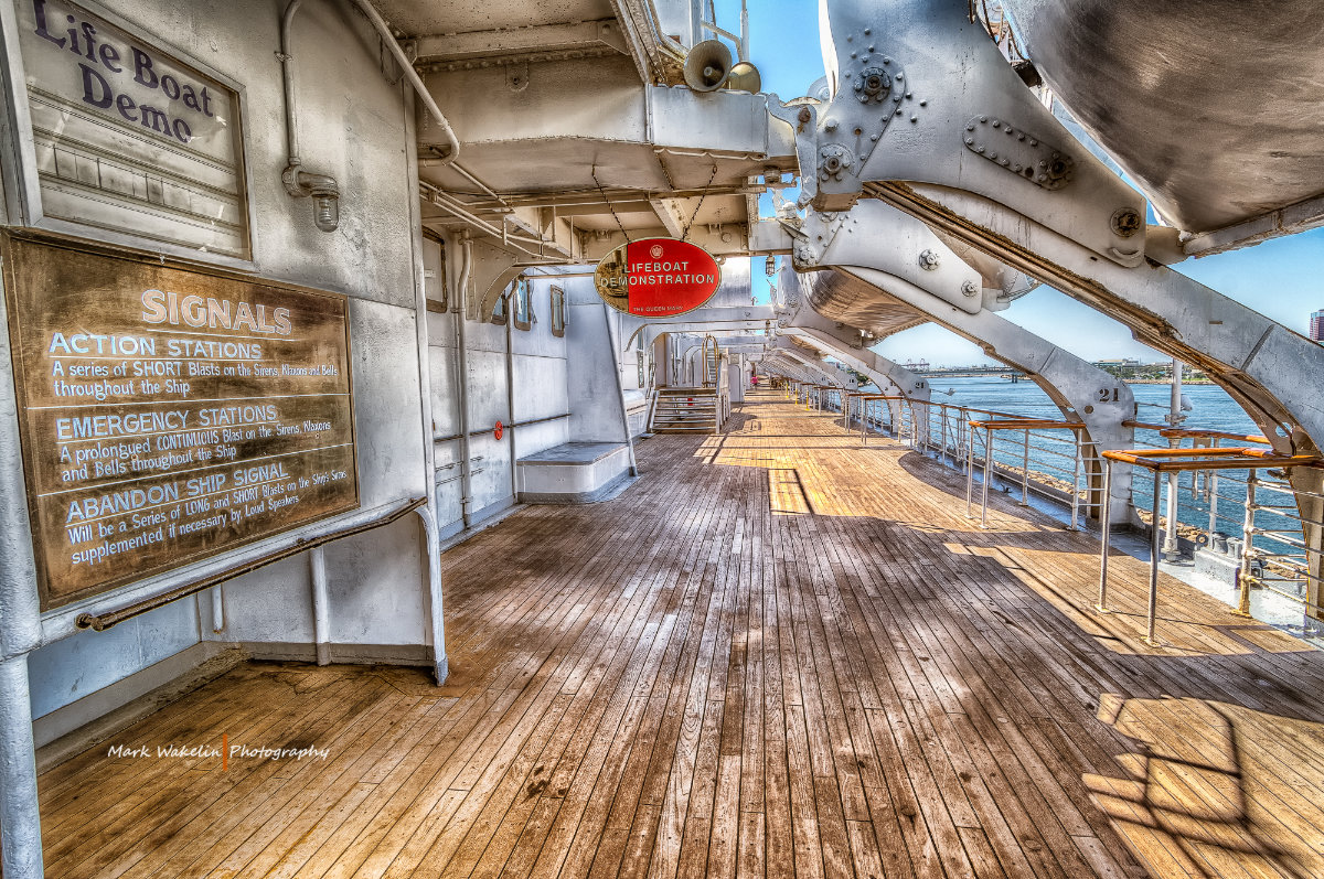 Wooden deck of a ship featuring lifeboat demonstration signage and metal lifeboat davits overlooking a calm body of water.