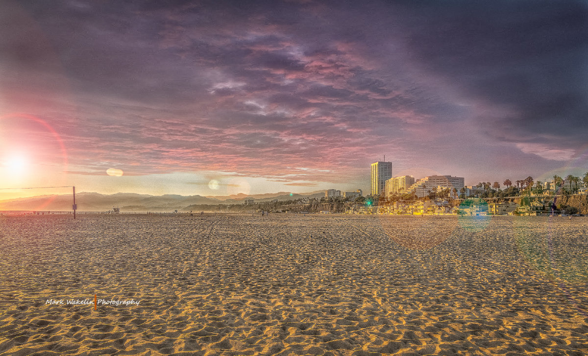 Wide sandy beach at sunset with coastal buildings and palm trees under a purple and pink cloudy sky.