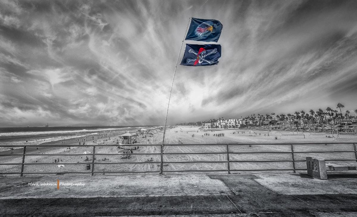 Two flags with eagle and pirate skull motifs flying from a pole on a pier overlooking a busy sandy beach under dramatic cloudy skies.