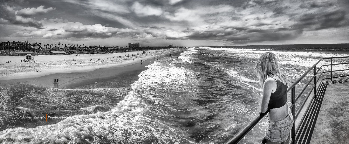 Black and white panoramic view of a beach with waves and scattered people, and a woman leaning on a railing looking at the sea.