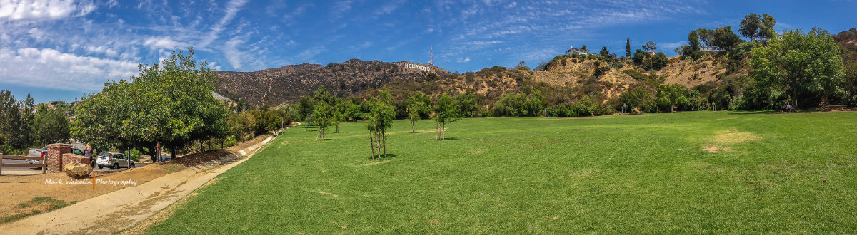 Panoramic view of a green park with small trees and the Hollywood sign visible on the hills under a partly cloudy blue sky.