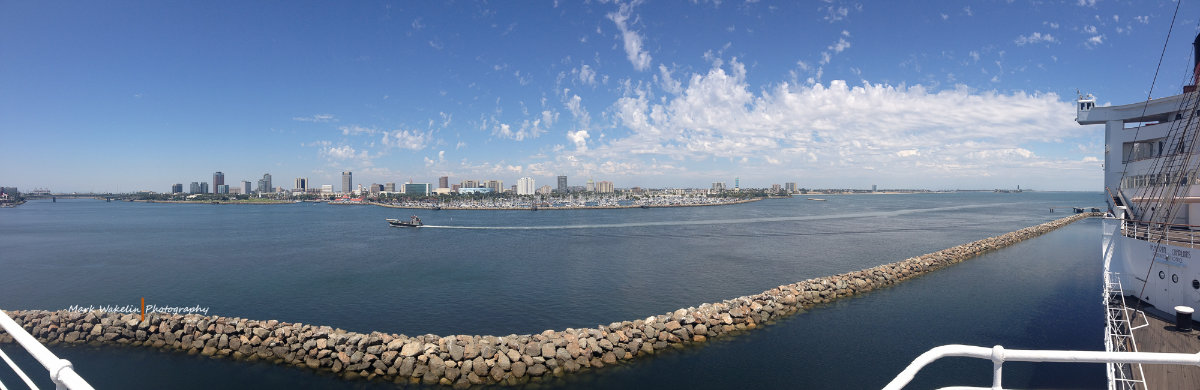 Wide panoramic view of a coastal city skyline with calm blue water, a breakwater, and a small boat leaving a wake under a partly cloudy sky.