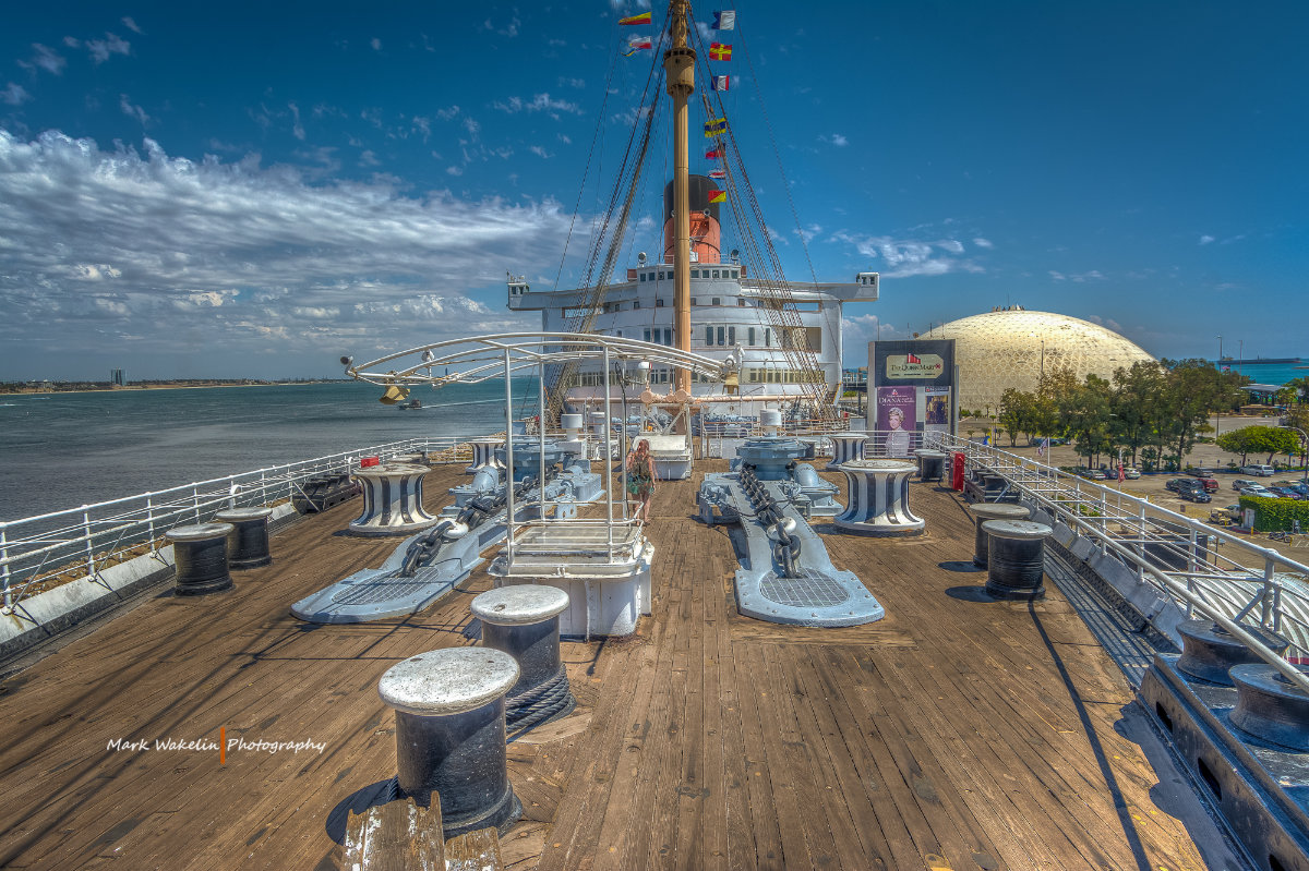 View from the wooden deck of a large ship with ropes, winches and chains, overlooking a river and a domed building under a partly cloudy blue sky.