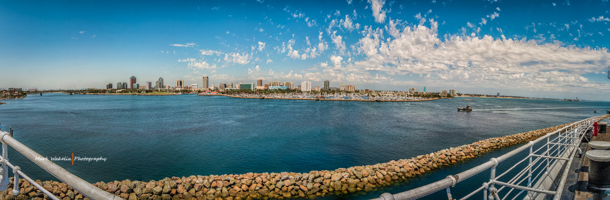Panoramic view of a large harbour with a city skyline and boats across the water under a partly cloudy blue sky.