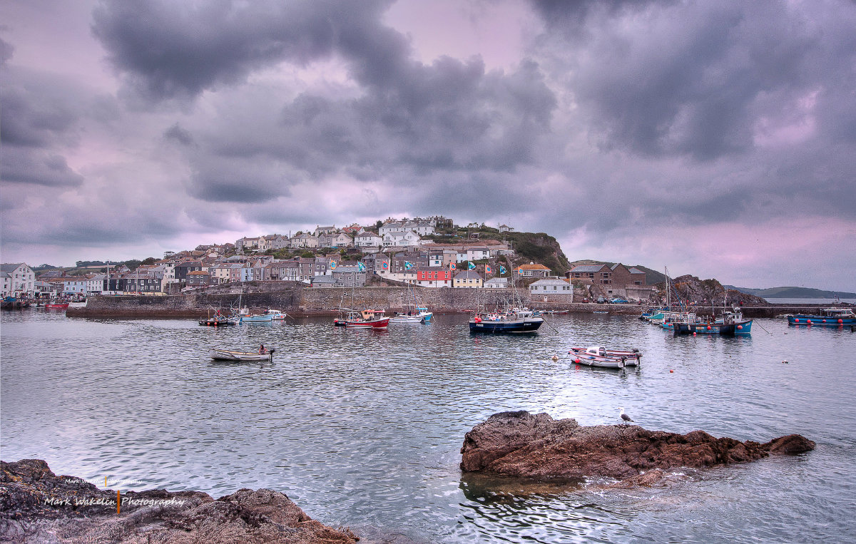 Fishing boats anchored in calm harbour waters with a hillside village under a cloudy sky.