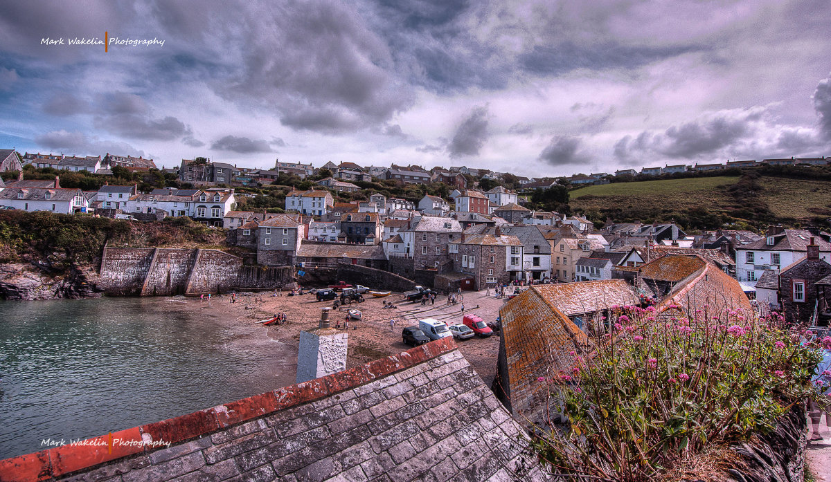 View over a coastal village with stone houses, a small harbour beach with boats and people, under a cloudy sky.