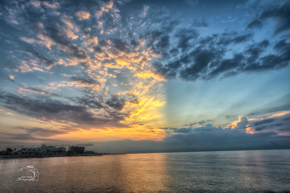 Sunset over calm sea with scattered clouds and buildings along the shoreline on the left.