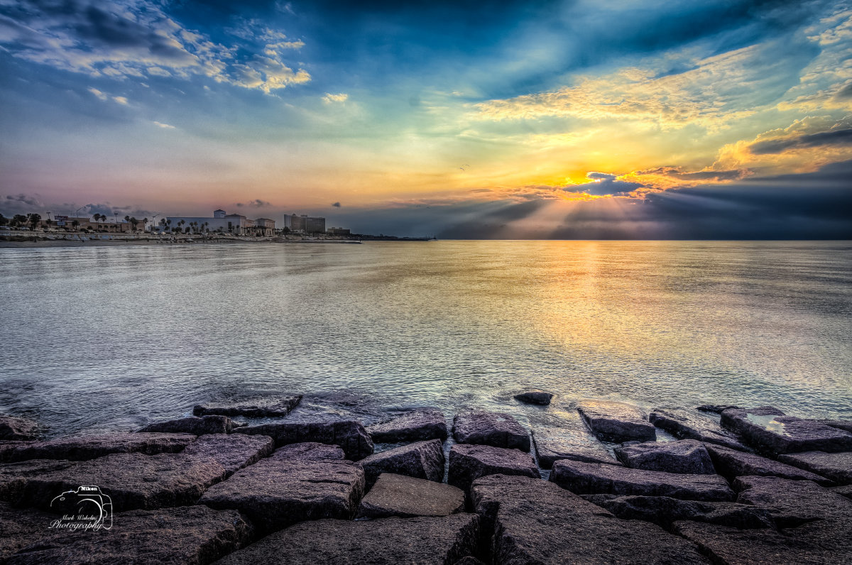 Sunset over calm sea with sun rays emerging through clouds, rocky shoreline in foreground and distant buildings along the coast.