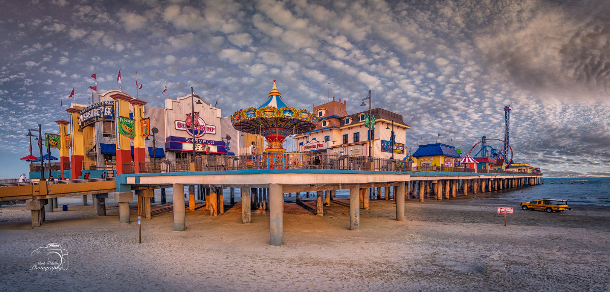 Wide view of a colourful seaside amusement pier with a carousel, shops, and rides under a cloudy sky at dusk.