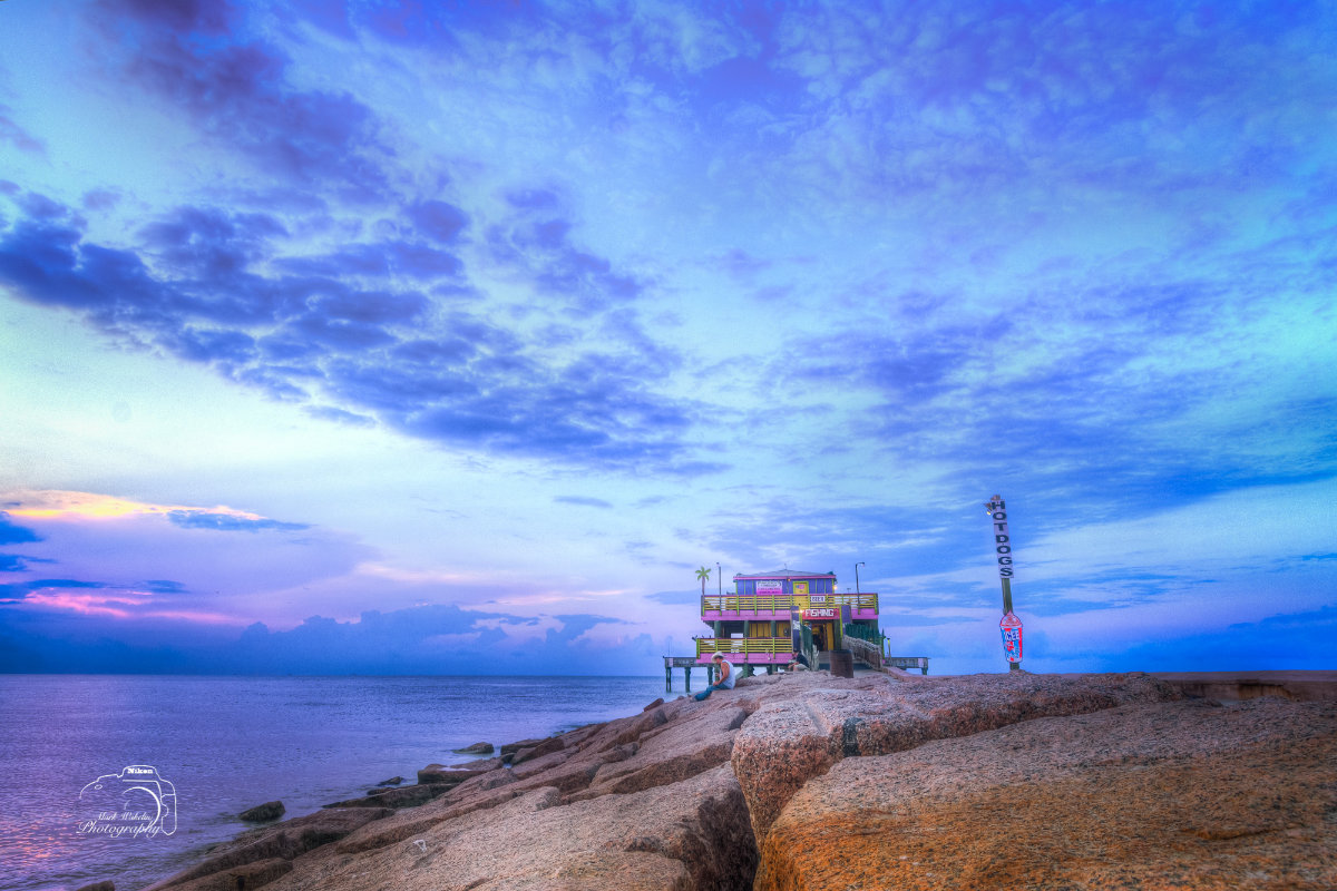 Seaside building on stilts at the end of rocky pier under a cloudy twilight sky.