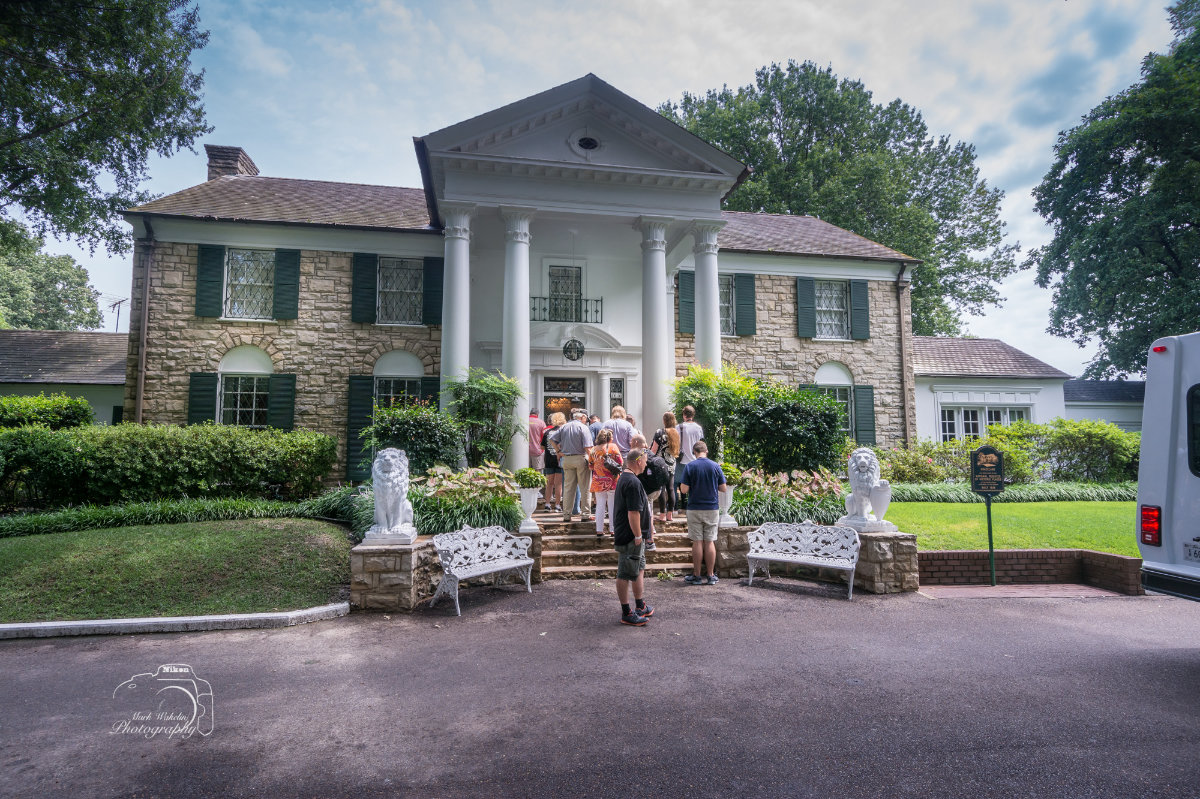 Group of people entering a stately stone house with white columns and green shutters, surrounded by greenery and two white lion statues.