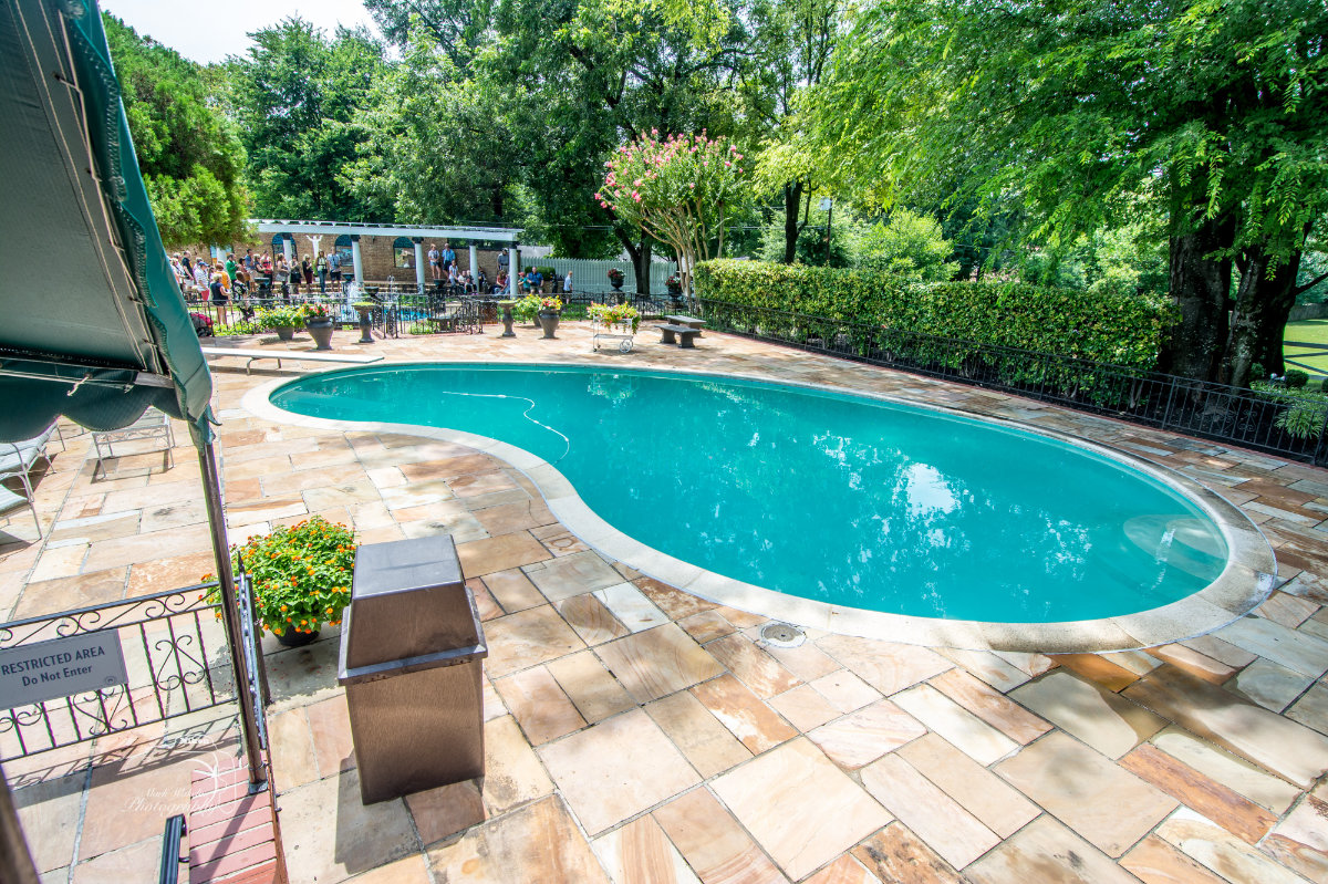 Kidney-shaped outdoor swimming pool surrounded by stone tiles, with lush green trees and a group of people gathered near a white pergola in the background.