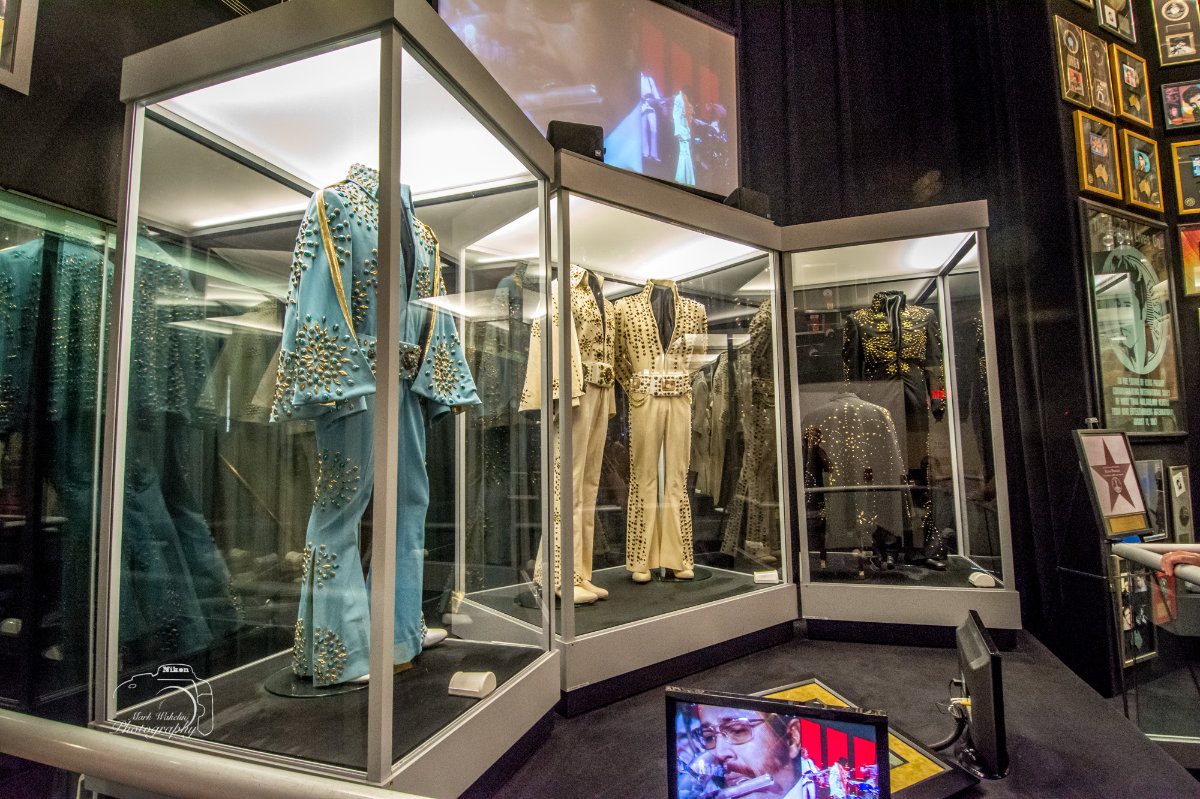 Museum display of vintage stage costumes with intricate studded designs in glass cases and a screen showing a musical performance.