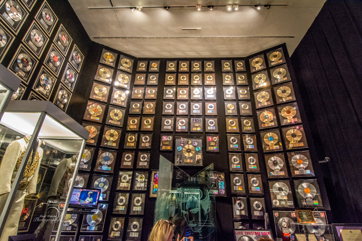 Wall covered with framed gold and platinum music awards in a museum display, with a glass case containing a white and gold costume to the left.