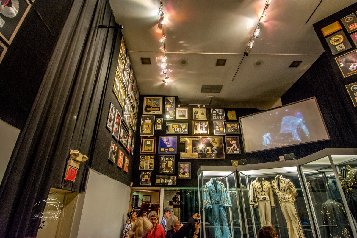 Museum room with Elvis Presley memorabilia including framed awards, a video screen showing a performance, and glass cases displaying elaborate stage costumes.