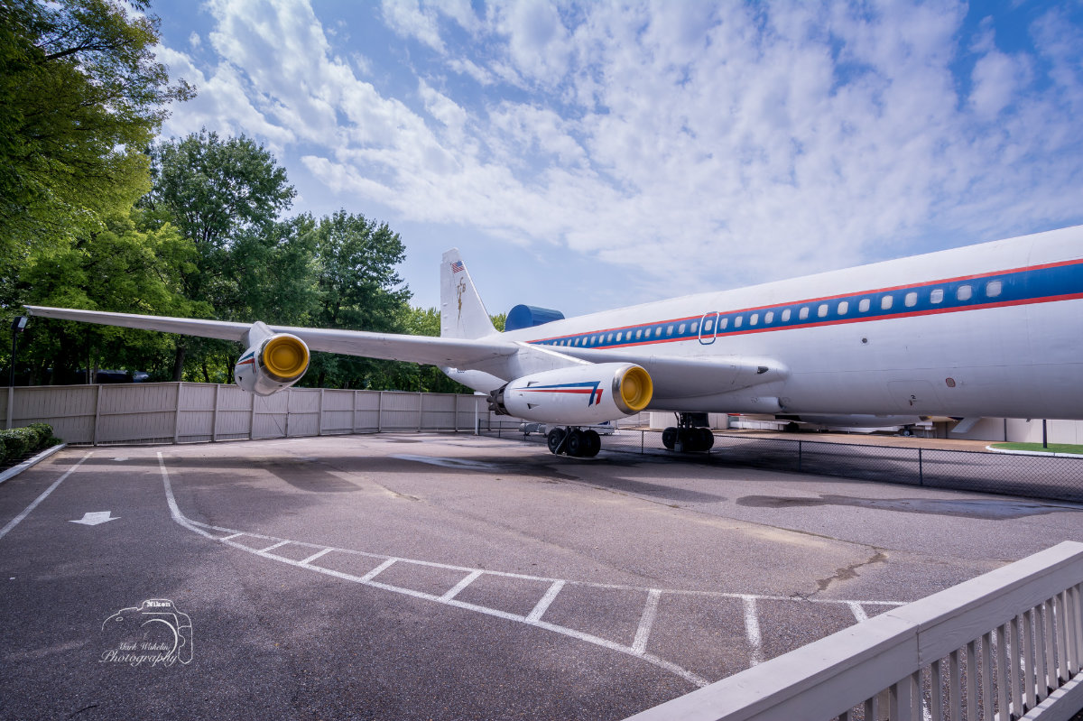 Rear view of a white vintage airplane with red and blue stripe, parked on tarmac beside a wooden fence and trees under a partly cloudy sky.