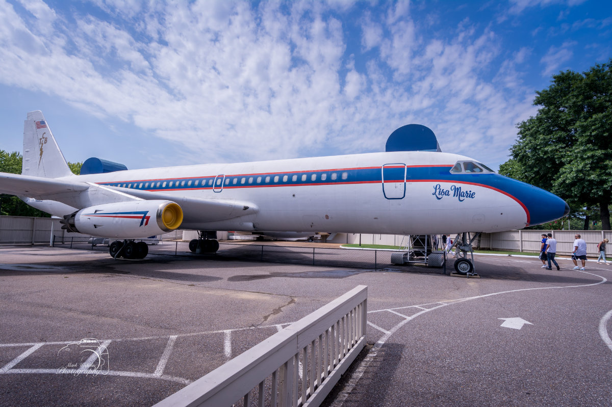 White vintage airplane named Lisa Marie with blue and red stripes parked outdoors on tarmac under a partly cloudy sky.