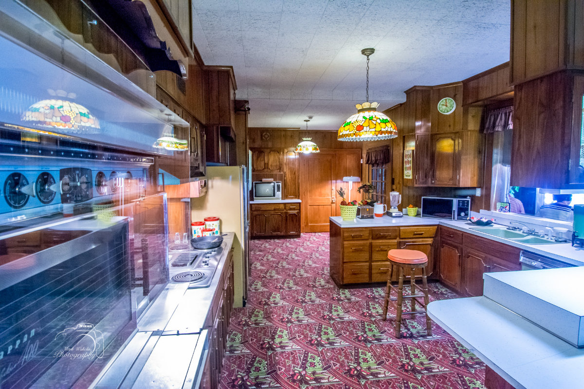 Vintage kitchen with wooden cabinets, patterned red carpet, stained glass pendant lights, and retro appliances.