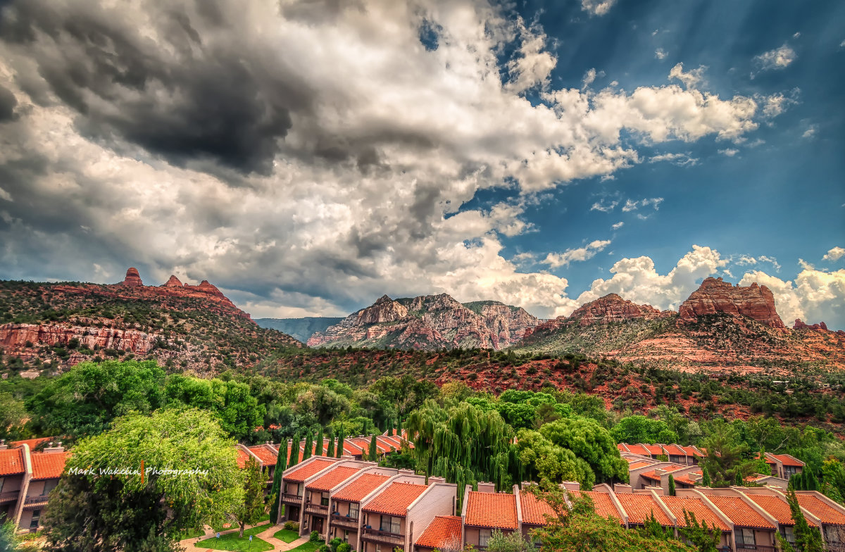 Red-roofed buildings surrounded by green trees with red rock mountains and a partly cloudy sky in the background.