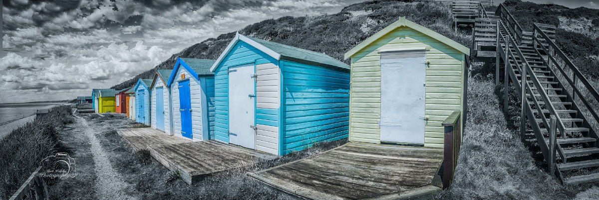 Line of colourful beach huts along a coastal path with wooden stairs on a grassy hill nearby.