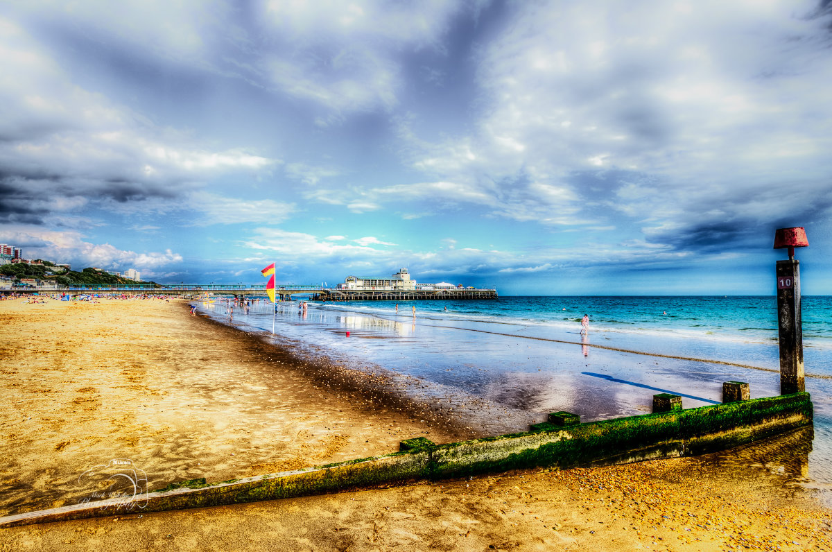 Sandy beach with people near the water, a pier extending into the sea under a partly cloudy sky, and a wooden groyne covered in moss in the foreground.