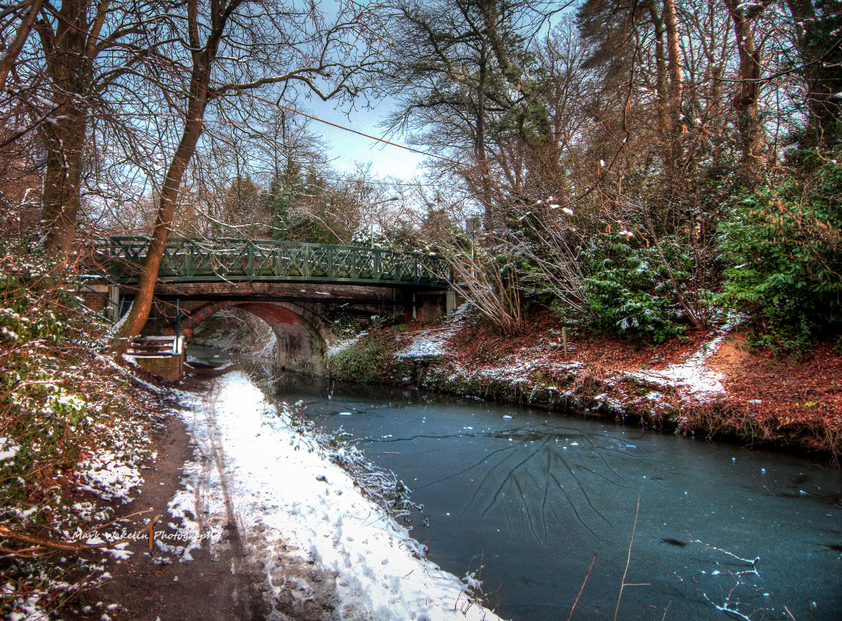 Snow-dusted riverside path next to a frozen river under a green metal bridge in a winter woodland.