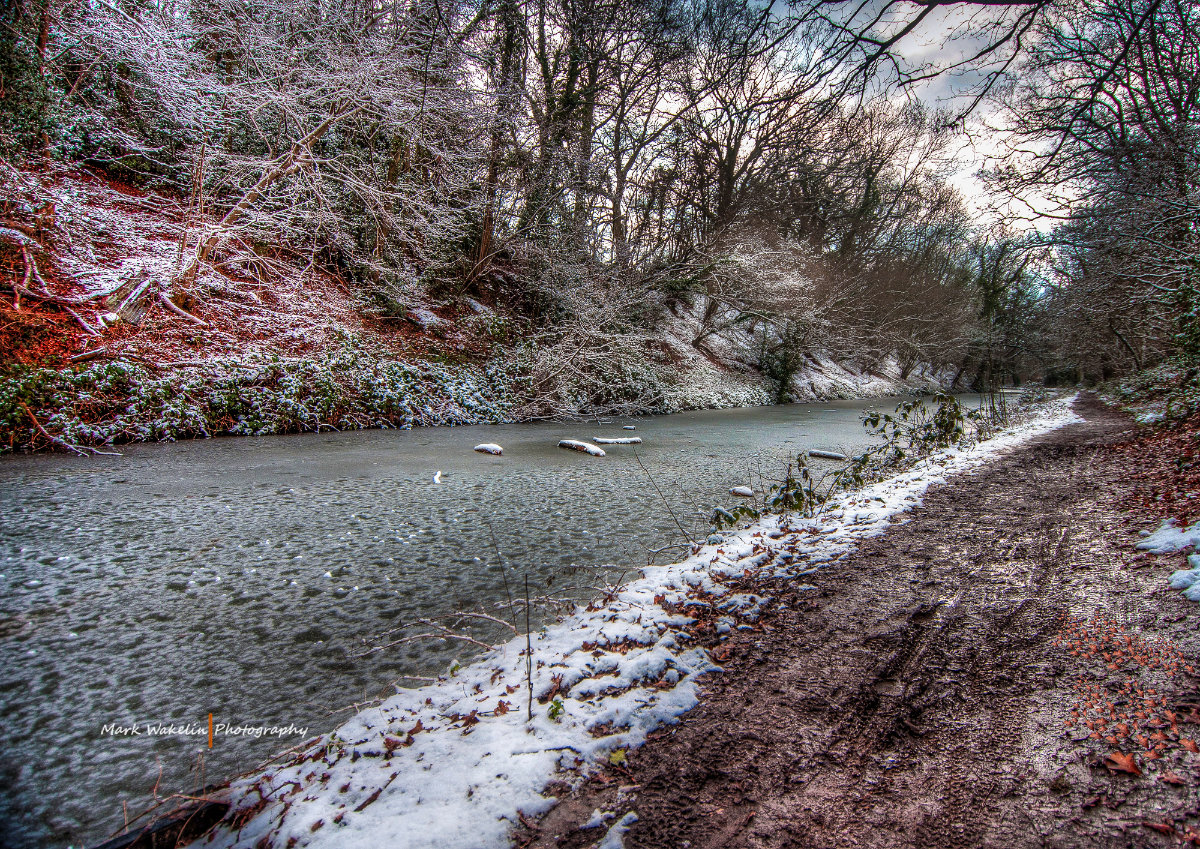 Snow-dusted trees and muddy footpath alongside a partially frozen river in winter.