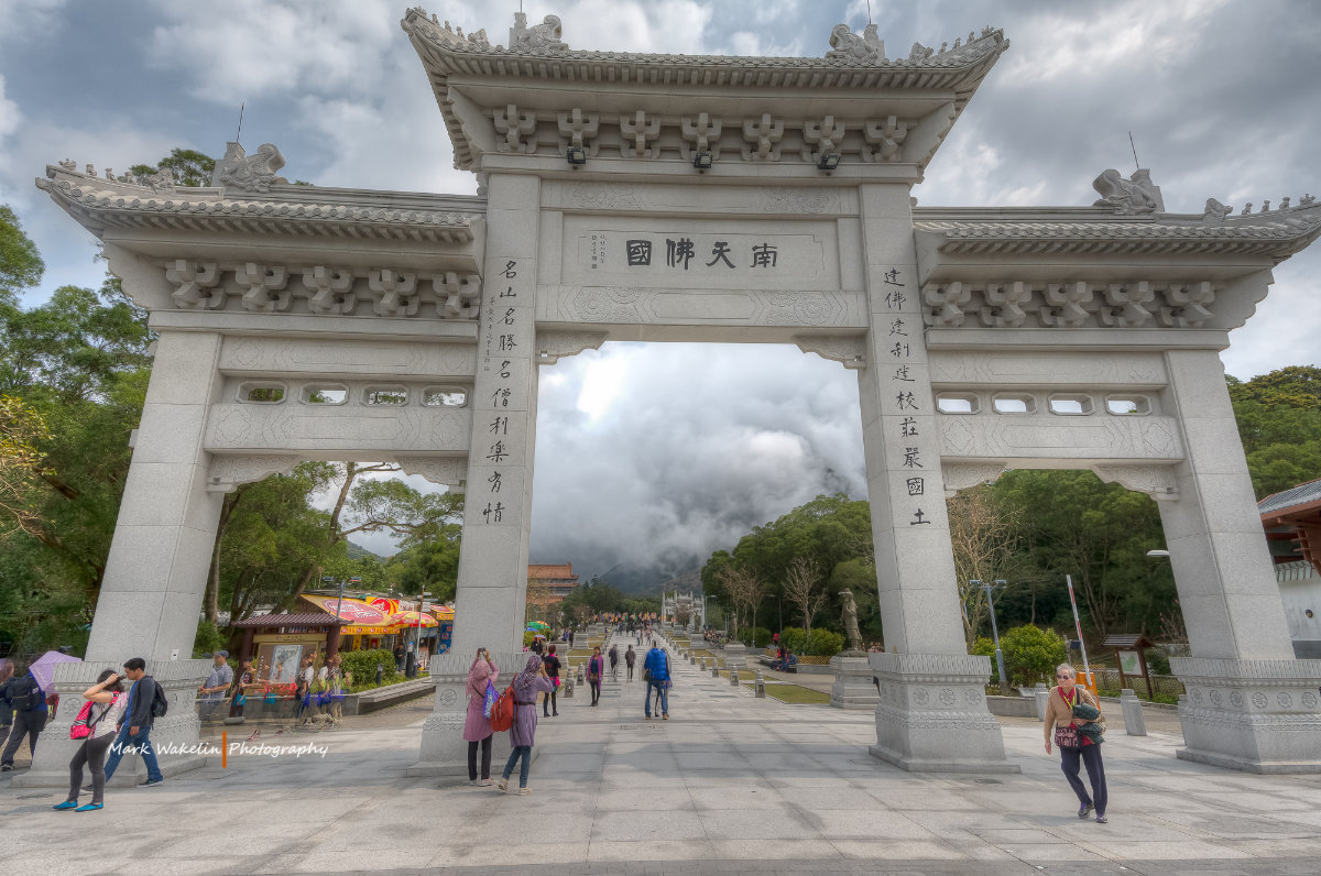 Large traditional Chinese stone archway with engraved calligraphy at the entrance to a park with people walking and trees under a cloudy sky.