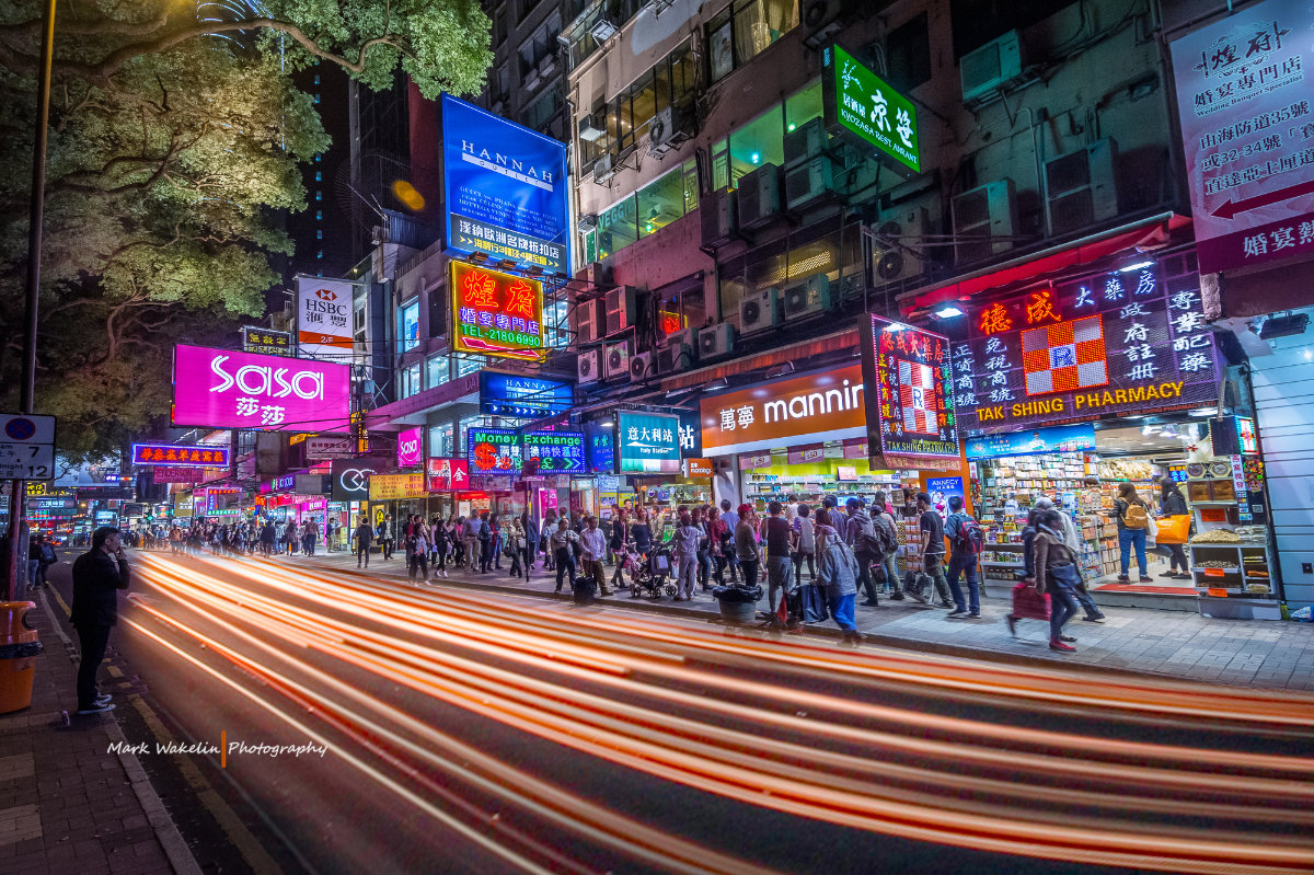 Busy night street scene in Hong Kong with vibrant neon signs, a crowd on the sidewalk, and light trails from passing vehicles.