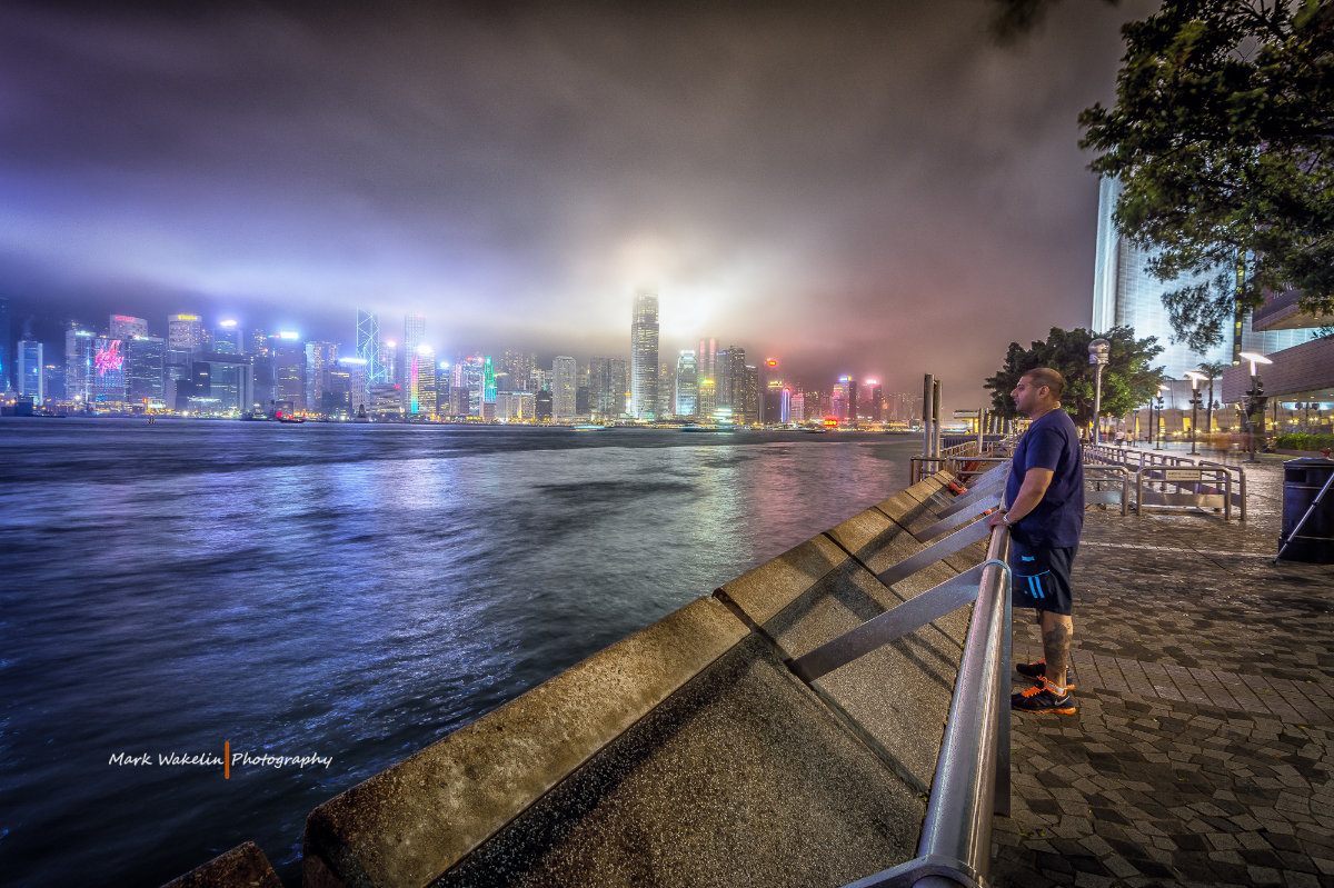 Man in casual clothes standing by a waterfront railing at night, looking across the water towards a brightly lit city skyline partially obscured by fog.