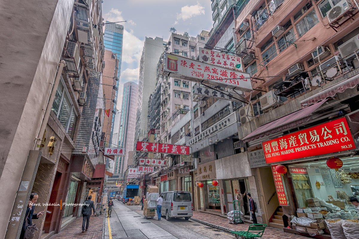 Narrow busy street lined with high-rise buildings featuring many hanging signs with Chinese characters, small shops, and people walking or working.