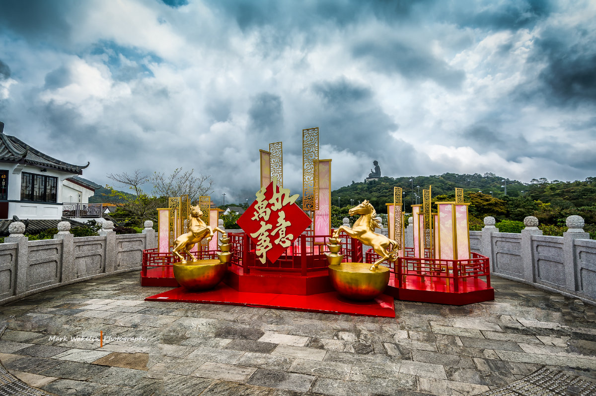 Golden horse statues on red platform with decorative panels and Chinese characters, overlooking the Tian Tan Buddha statue on a hillside under a cloudy sky.