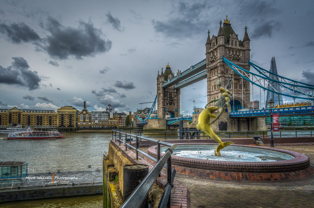 View of Tower Bridge over the River Thames with the Girl with a Dolphin statue in the foreground and The Shard visible in the background under a cloudy sky.