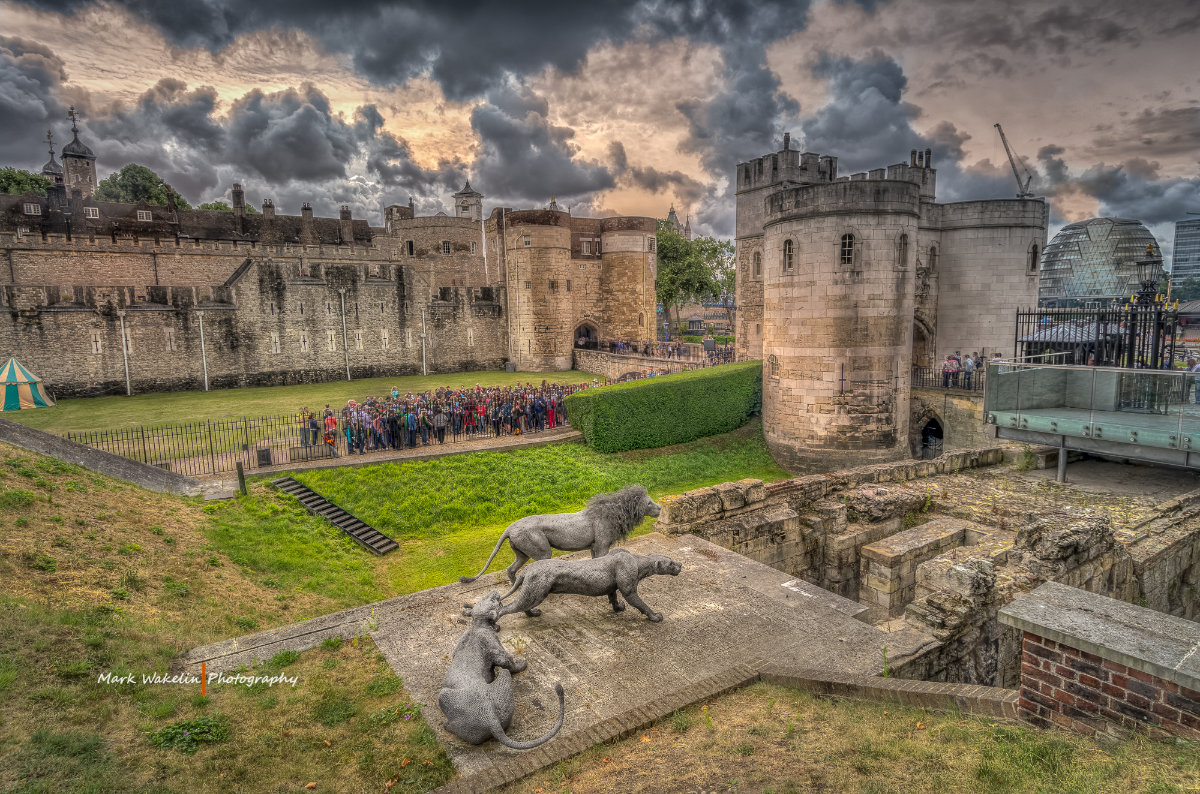 Stone sculptures of three lions overlooking the moat and entrance of the Tower of London under a cloudy sky.