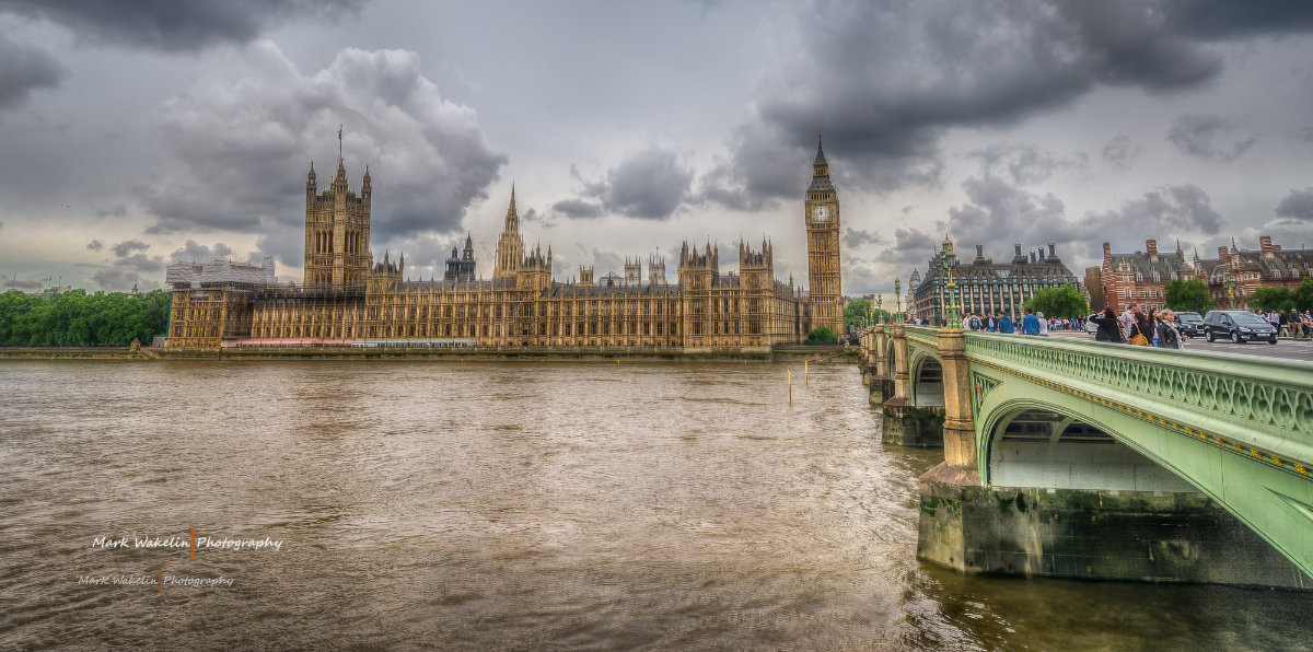 View of the Houses of Parliament and Big Ben beside the River Thames under a cloudy sky, with Westminster Bridge on the right busy with pedestrians and cars.