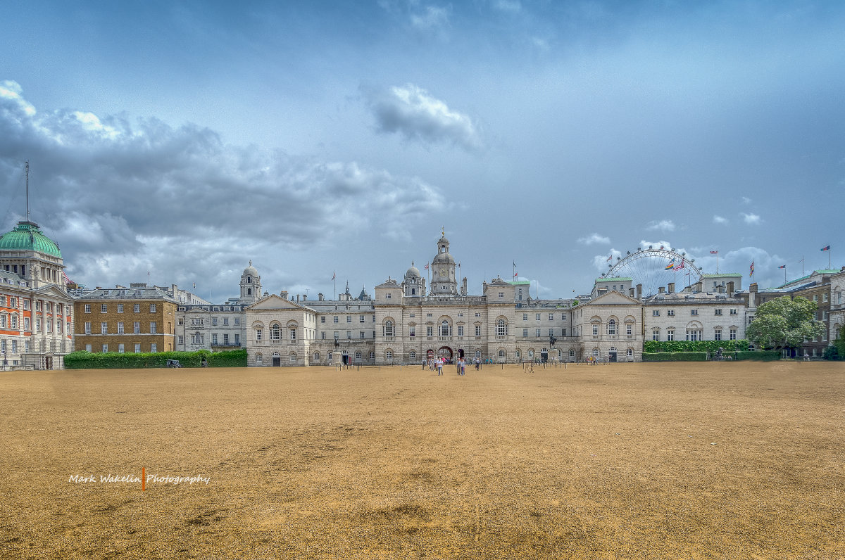 Historic grand building with light stone façade and central clock tower under a cloudy sky, viewed across a large dry grass field.
