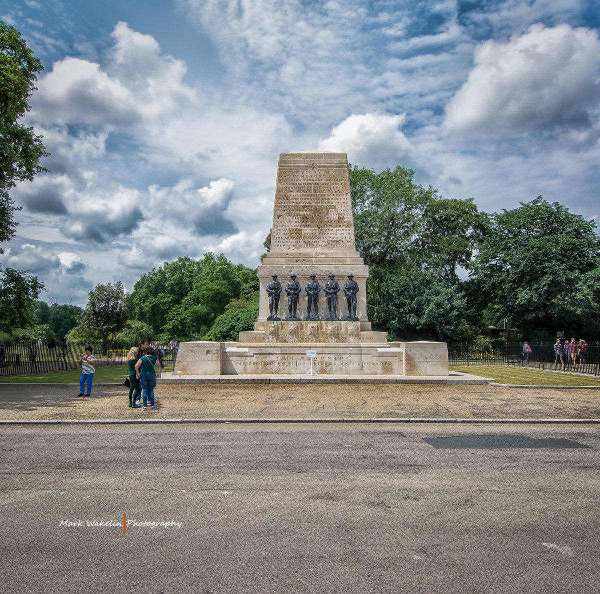 Stone war memorial monument with five soldier statues in front, set in a park with trees under a partly cloudy sky.