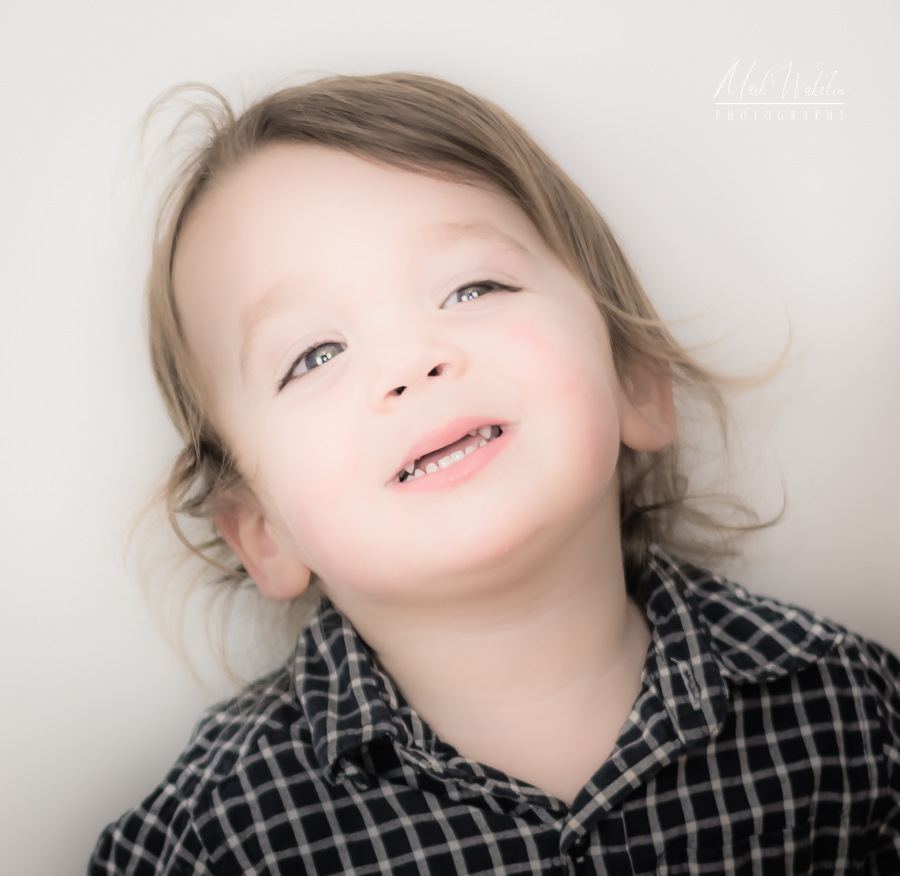 Toddler with light brown hair wearing a black and white checkered shirt smiling against a plain white background.