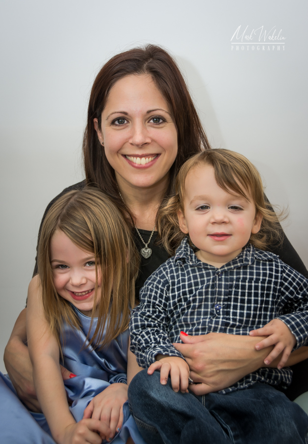 Smiling woman with brown hair hugging two young children, a girl in a blue dress and a boy in a checkered shirt.