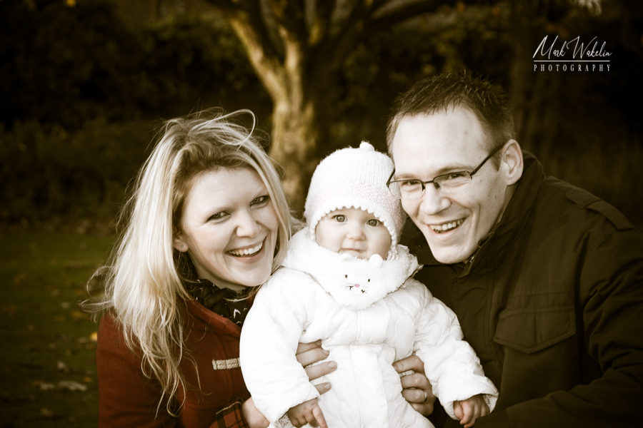 Smiling couple holding a baby dressed in a white coat and knit hat outdoors with trees in the background.