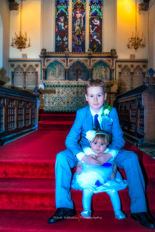 A boy in a blue suit sitting on red-carpeted church steps holding a baby girl in a white dress with a flower headband, in front of a decorated altar and stained glass windows.